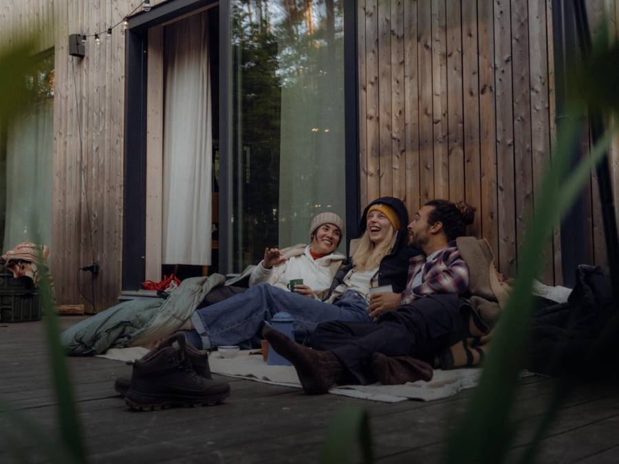 Trois personnes en tenue décontractée de plein air sont assises, blotties les unes contre les autres, sur la véranda d'une cabane en bois dans la forêt.